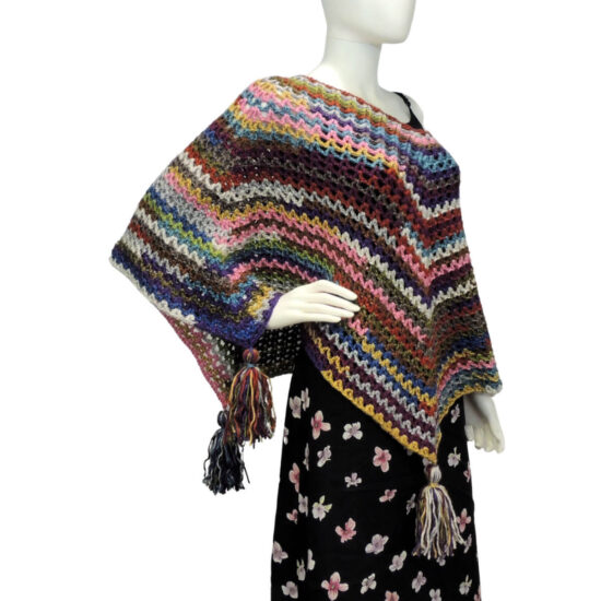 Front of a mannequin wearing a black skirt and a multicolor V-stitch crochet square poncho with large tassels hanging from each of the 4 points. The points hang on the two sides, with one of them draped over the mannequin's bent arm.