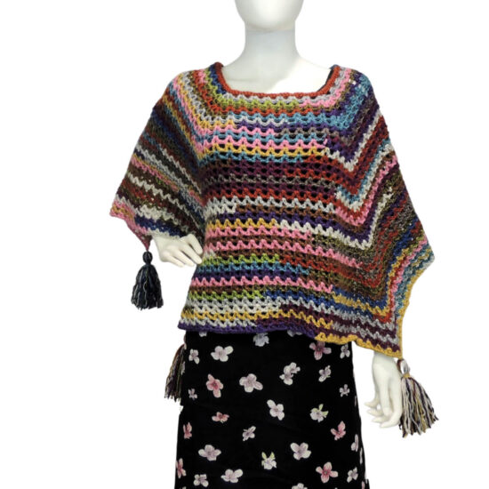 Front of a mannequin wearing a black skirt and a multicolor V-stitch crochet square poncho with large tassels hanging from each of the 4 points. The points hang on the two sides, with one of them draped over the mannequin's bent arm.
